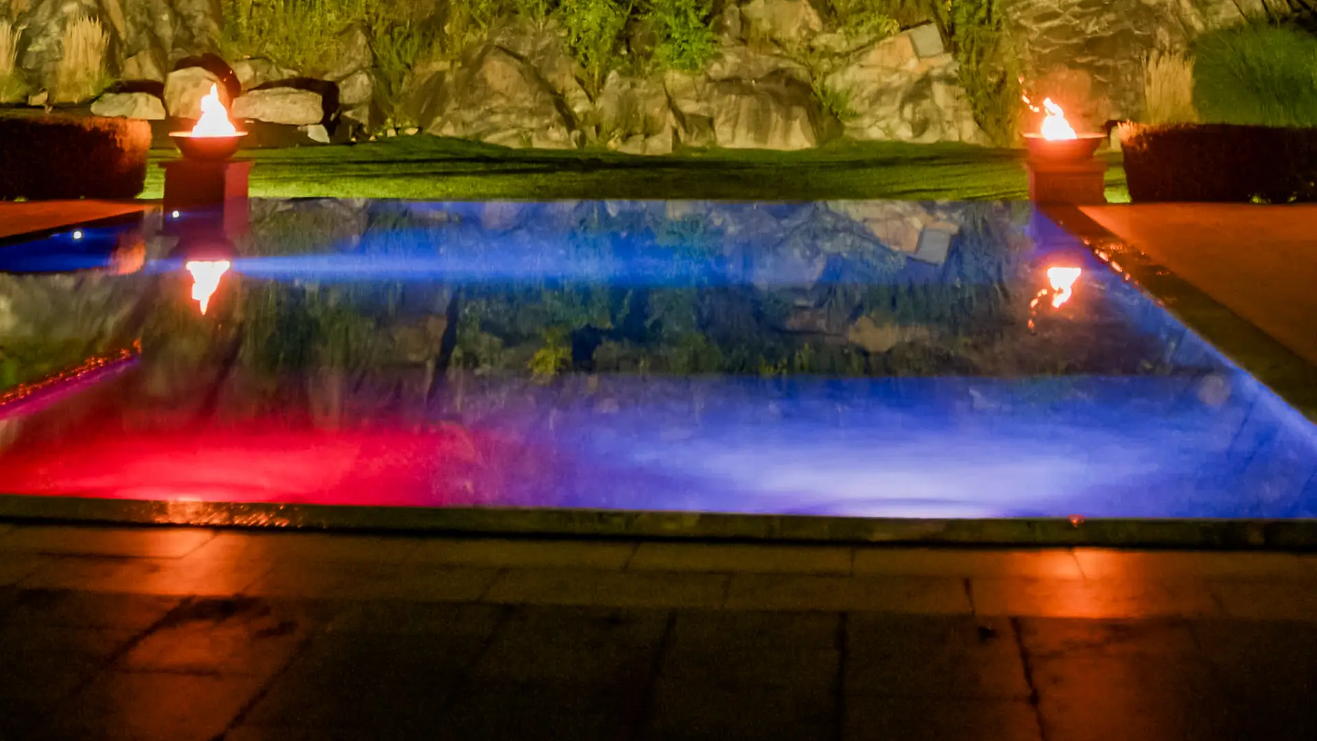 Rock-face pool with fire bowls lit at night, built by Artisan Landscapes and Pools